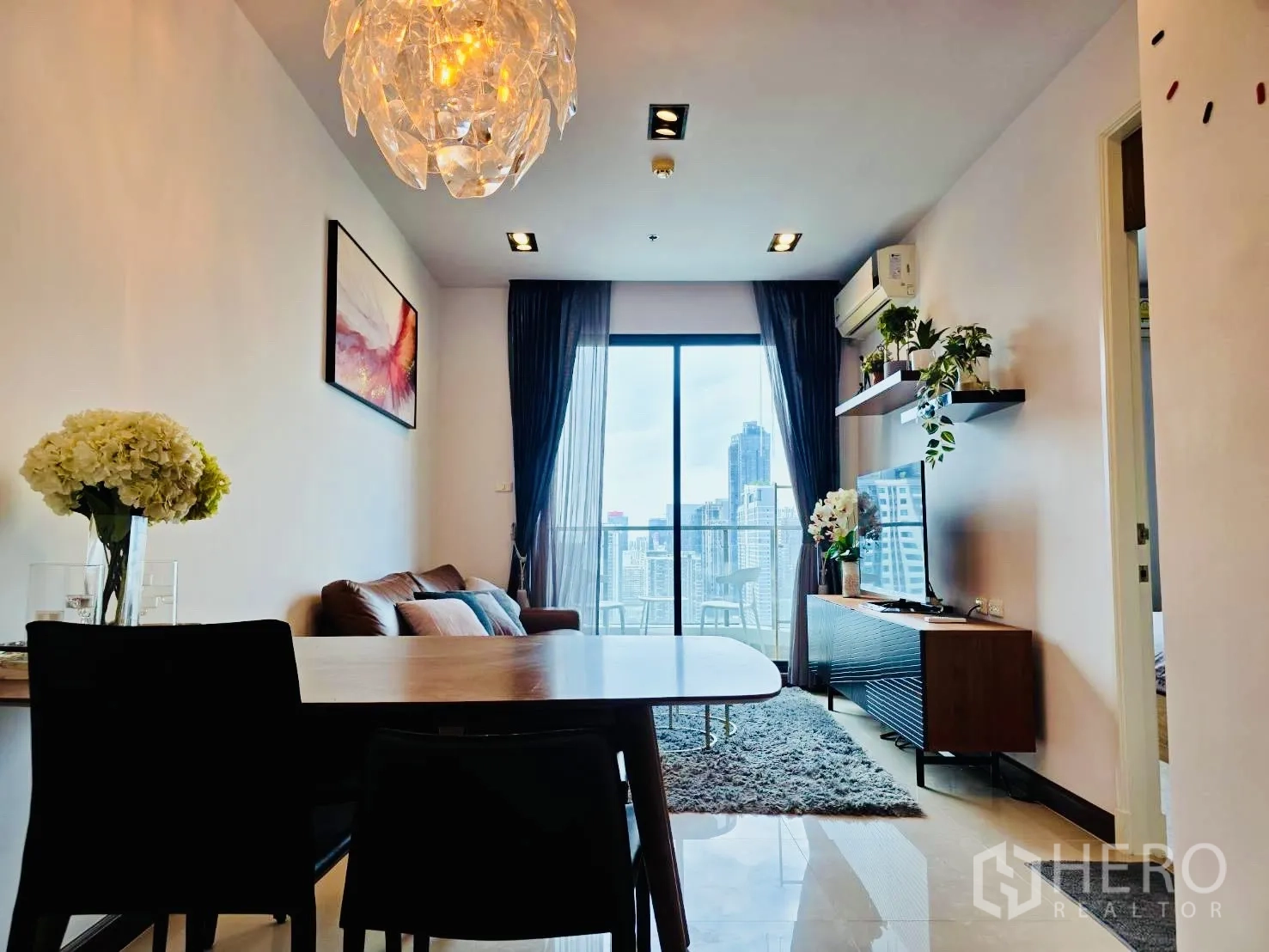 Condominium for rent in Huai Khwang, Bangkok - Dining table and sofa under a sculptural pendant beside floor-to-ceiling glass to the balcony and skyline.