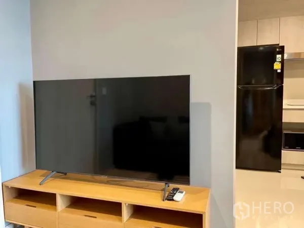 Condominium for rent in Pathum Wan, Bangkok - Flat-screen TV on wooden media unit beside entrance to compact kitchen with refrigerator