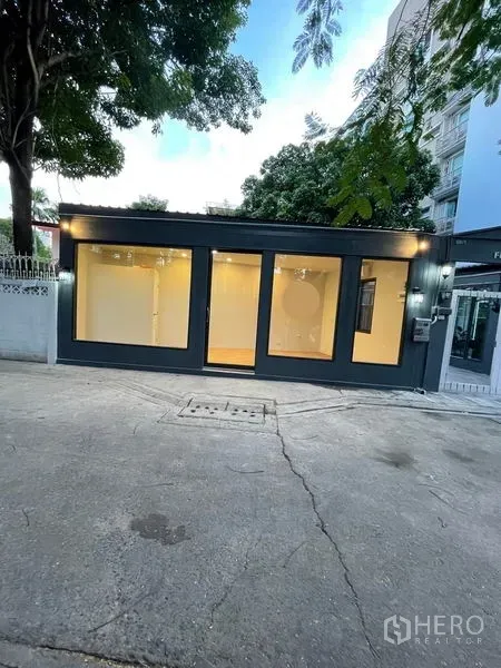 พื้นที่ขายปลีก ให้เช่าในKhlong Toei, Bangkok - อาคารร้านค้าชั้นเดียวริมถนน โครงสีเข้ม หน้ากระจกเต็มบาน ภายในไฟอบอุ่น มีต้นไม้และอาคารพักอาศัยย่านคลองเตย กรุงเทพฯ