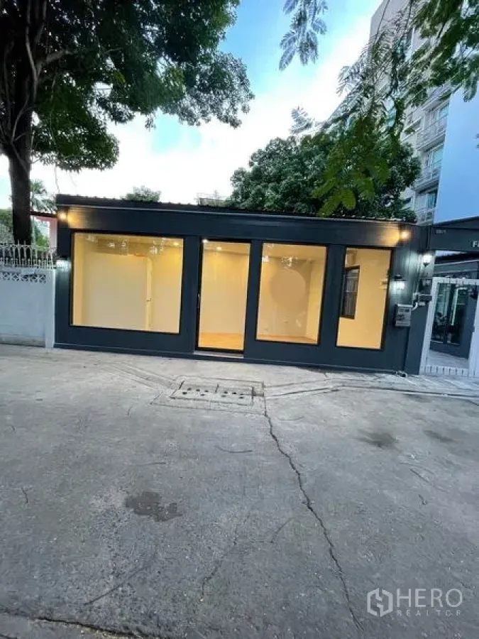 พื้นที่ขายปลีก ให้เช่าในKhlong Toei, Bangkok - อาคารร้านค้าชั้นเดียวริมถนน โครงสีเข้ม หน้ากระจกเต็มบาน ภายในไฟอบอุ่น มีต้นไม้และอาคารพักอาศัยย่านคลองเตย กรุงเทพฯ