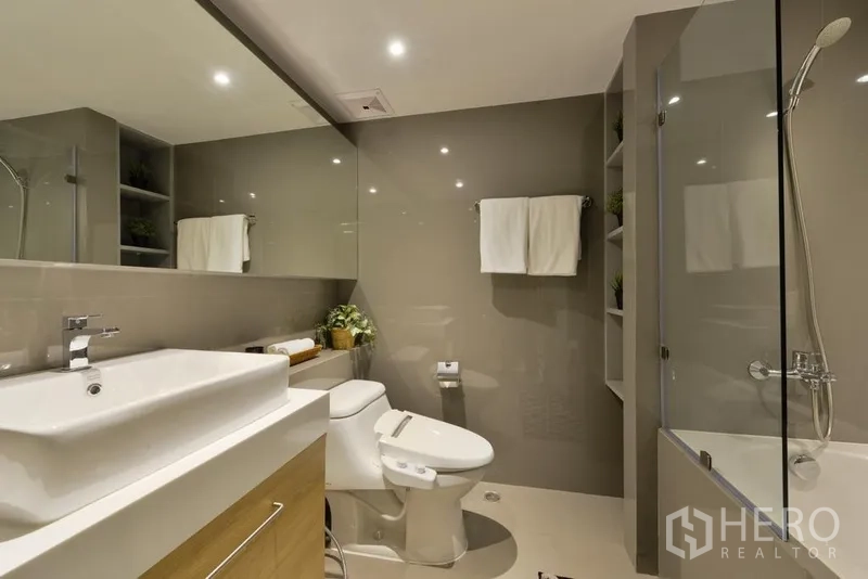 Apartment for rent in Khlong Toei, Bangkok - Contemporary bathroom with wide vanity, large mirror, toilet and glass shower screen.