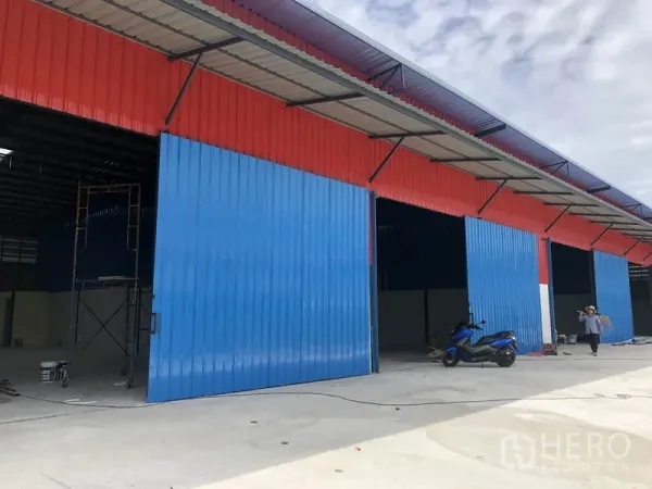 Warehouse for rent in Khlong Luang, Pathum Thani - Row of new warehouses with red cladding and large blue sliding steel doors, wide concrete driveway and covered awnings.