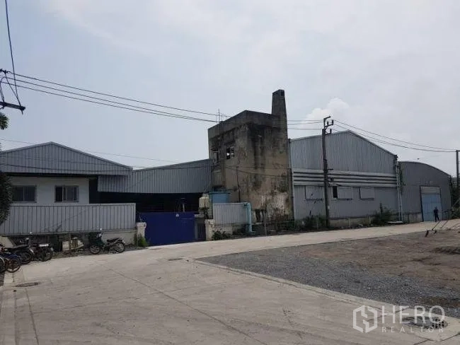 Factory for rent or sale in Bang Phli, Samut Prakan - Wide concrete access road leading to clustered metal factory buildings with a tall concrete stack.