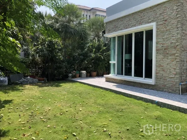 Detached House for rent or sale in Saphan Sung, Bangkok - Private landscaped backyard with green lawn, trees, and a stone-clad exterior wall with large sliding doors.
