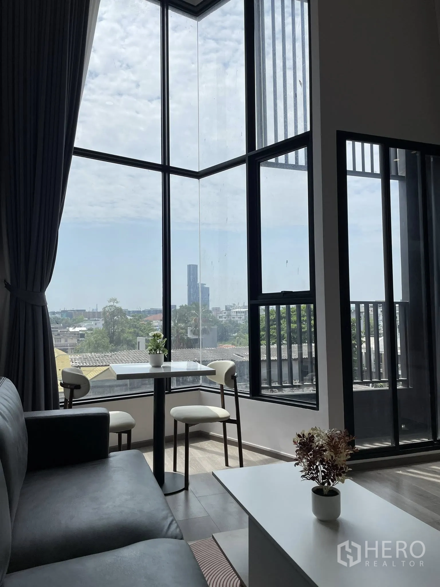 Condominium for rent in Khlong Toei, Bangkok - Bright living area with double-height corner windows, a small dining set, and city views.