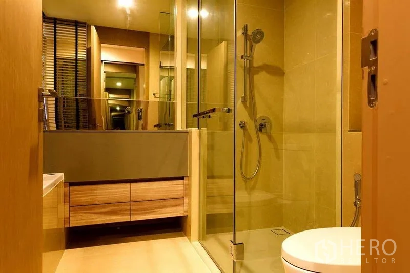 Condominium for rent or sale in Watthana, Bangkok - Warm-toned bathroom with glass shower enclosure, rain shower and wood vanity.