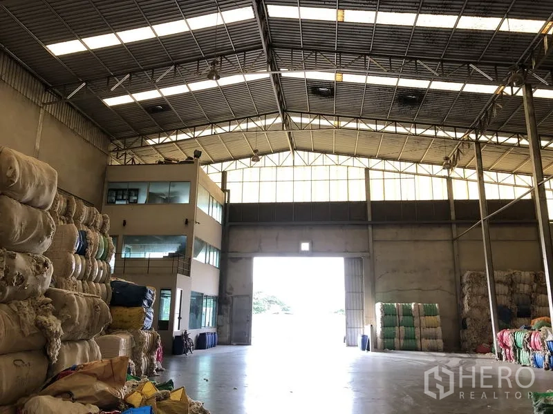 工厂 出租在Bang Bua Thong, Nonthaburi - 仓库入口处高大卷门，旁接带玻璃窗的办公室楼。