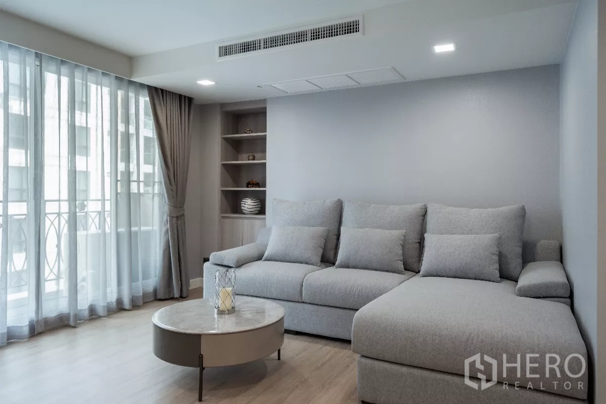 Apartment for rent - Bright living room with gray L-shaped sofa, sheer curtained balcony, built-in shelving and round marble coffee table.