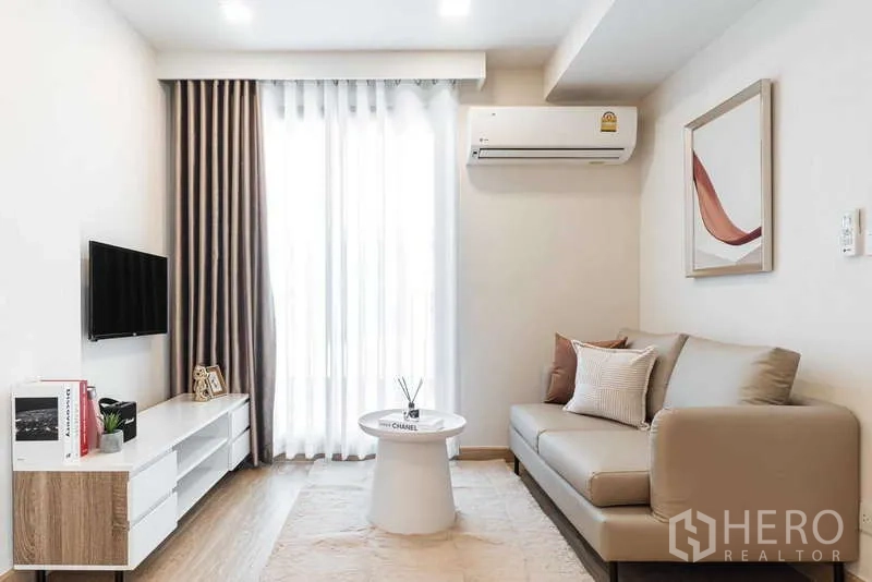 คอนโด ให้เช่า - ห้องนั่งเล่นพร้อมชั้นวางทีวี เครื่องปรับอากาศ และผ้าม่านโปร่งสู่ระเบียง