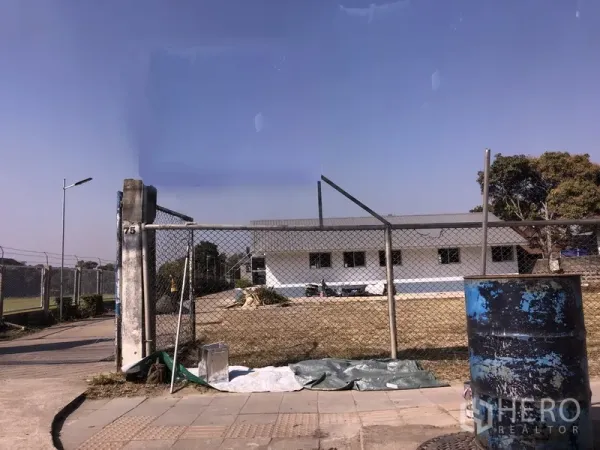 Warehouse for rent in Pathum Thani - Gated industrial compound with chain-link fence, open yard, and a low white warehouse building under a clear sky in Pathum Thani.