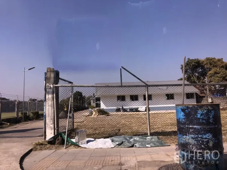 Warehouse for rent in Pathum Thani - Gated industrial compound with chain-link fence, open yard, and a low white warehouse building under a clear sky in Pathum Thani.