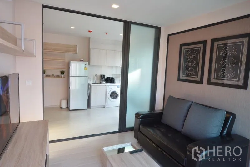 คอนโด ขายในKhlong Toei, Bangkok - ห้องนั่งเล่นสว่าง โซฟาหนังสีดำ ตู้ทีวี และประตูกระจกบานเลื่อนเชื่อมห้องครัวพร้อมตู้เย็นและเครื่องซักผ้า