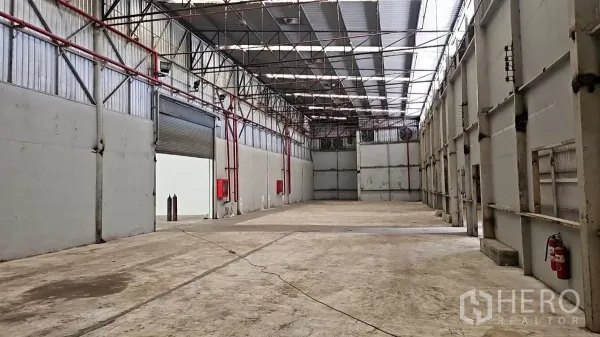 工厂 出租在Pak Kret, Nonthaburi - 宽敞厂房大厅，钢结构高挑屋顶带天窗，卷闸门与消防箱，地面为混凝土。
