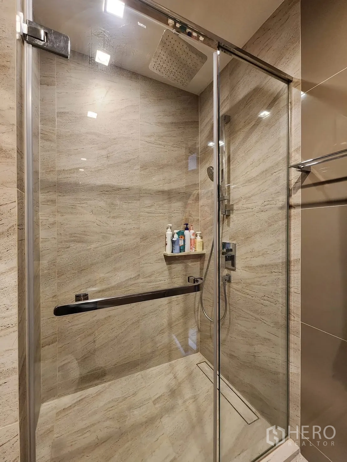 Condominium for rent in Watthana, Bangkok - Modern bathroom featuring a glass-enclosed rain shower with beige stone tiles and built-in shelf.