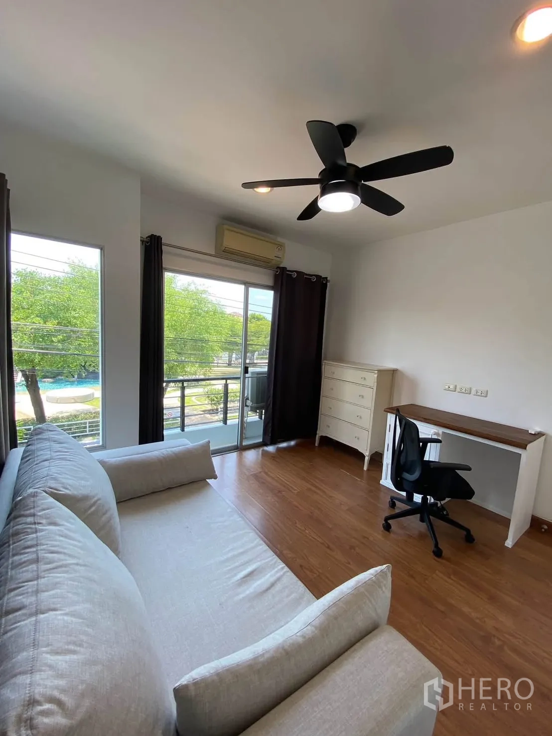 Detached House for rent in Mueang Samut Prakan, Samut Prakan - Bright room with sofa, desk, and balcony overlooking greenery and pool.