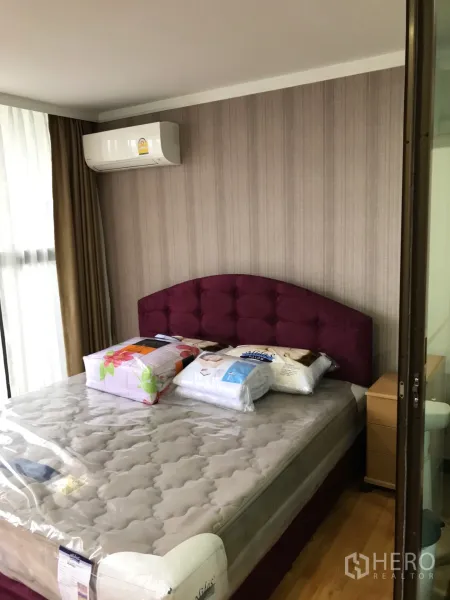 Condominium for rent or sale in Watthana, Bangkok - Bright bedroom with burgundy tufted headboard, air conditioner and floor‑to‑ceiling curtains in a Sukhumvit condo.