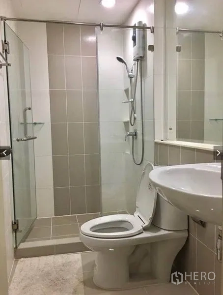Townhouse for rent or sale in Suan Luang, Bangkok - Bathroom with glass-enclosed shower, water heater, toilet, and wall-mounted sink in neutral tiles.