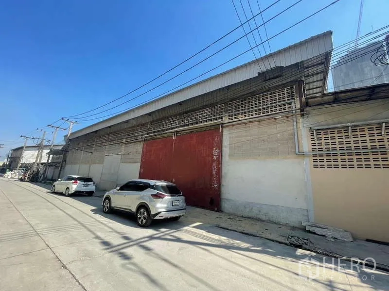Warehouse for rent in Rat Burana, Bangkok - Street-facing warehouse facade with large red metal door, high roof and roadside parking in the Rat Burana industrial area of Bangkok.