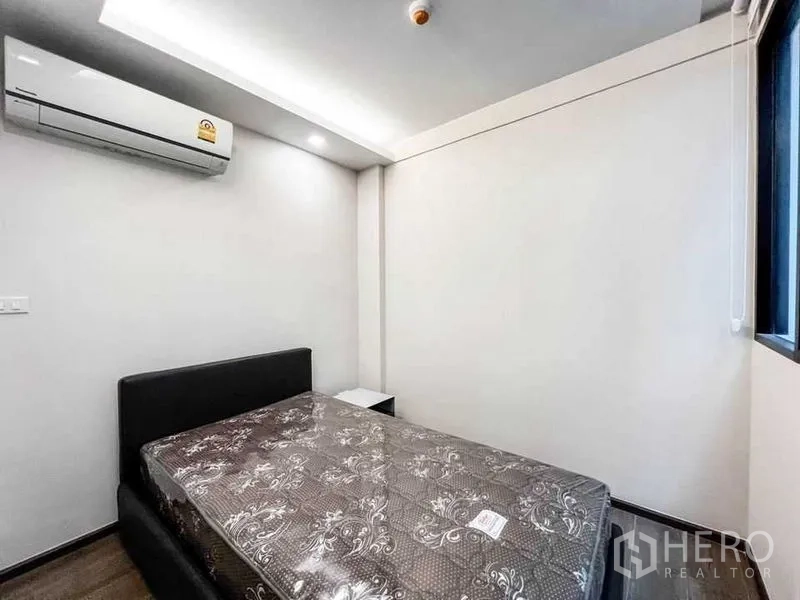 Condominium for sale in Huai Khwang, Bangkok - Compact bedroom with double bed, air conditioner, white walls and a window for natural light.