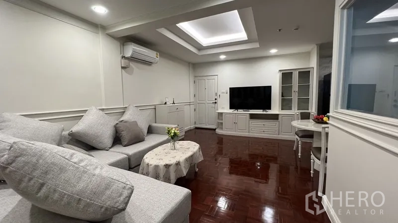 Apartment for rent in Watthana, Bangkok - Bright living room with L-shaped gray sofa, tray ceiling, TV console and glossy parquet floors.