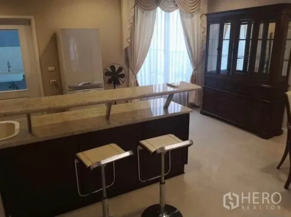 Detached House for rent in Wang Thonglang, Bangkok - Granite-topped kitchen island with two bar stools and a tall display cabinet by curtained window.
