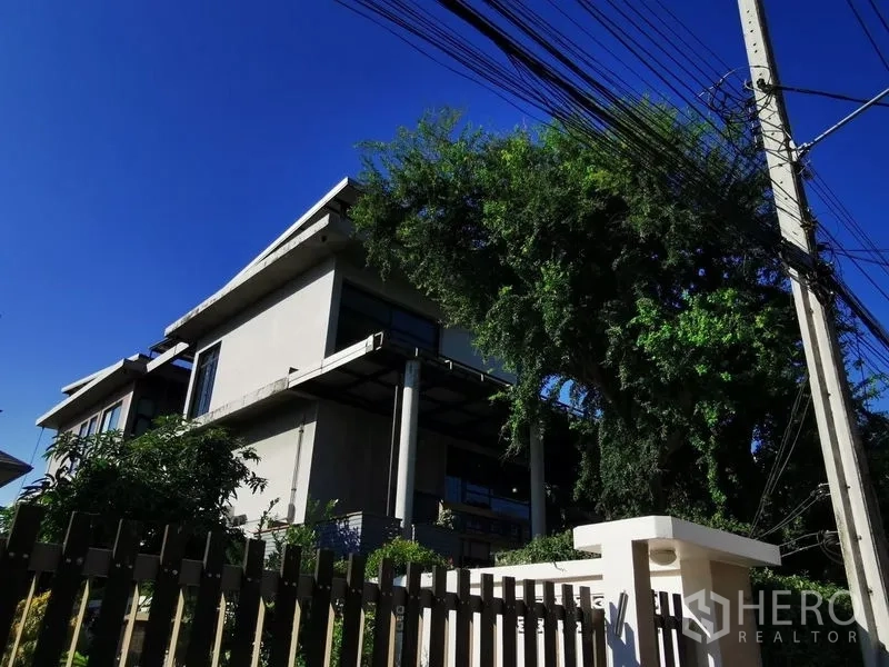 Home Office for sale in Wang Thonglang, Bangkok - Street view of a modern concrete building behind a fence and mature tree.