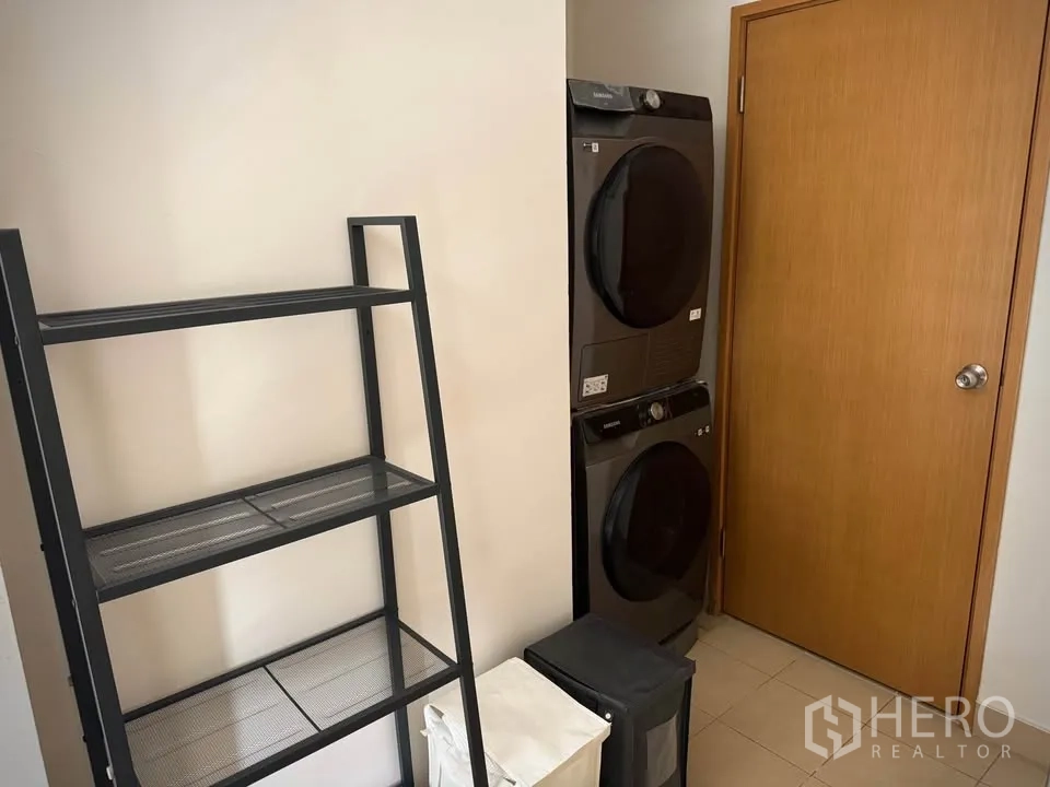 คอนโด for rent or sale in Khlong Toei, Bangkok - ห้องซักล้างพร้อมเครื่องซักและอบผ้าแบบวางซ้อน ชั้นวางโลหะ และประตูไม้.