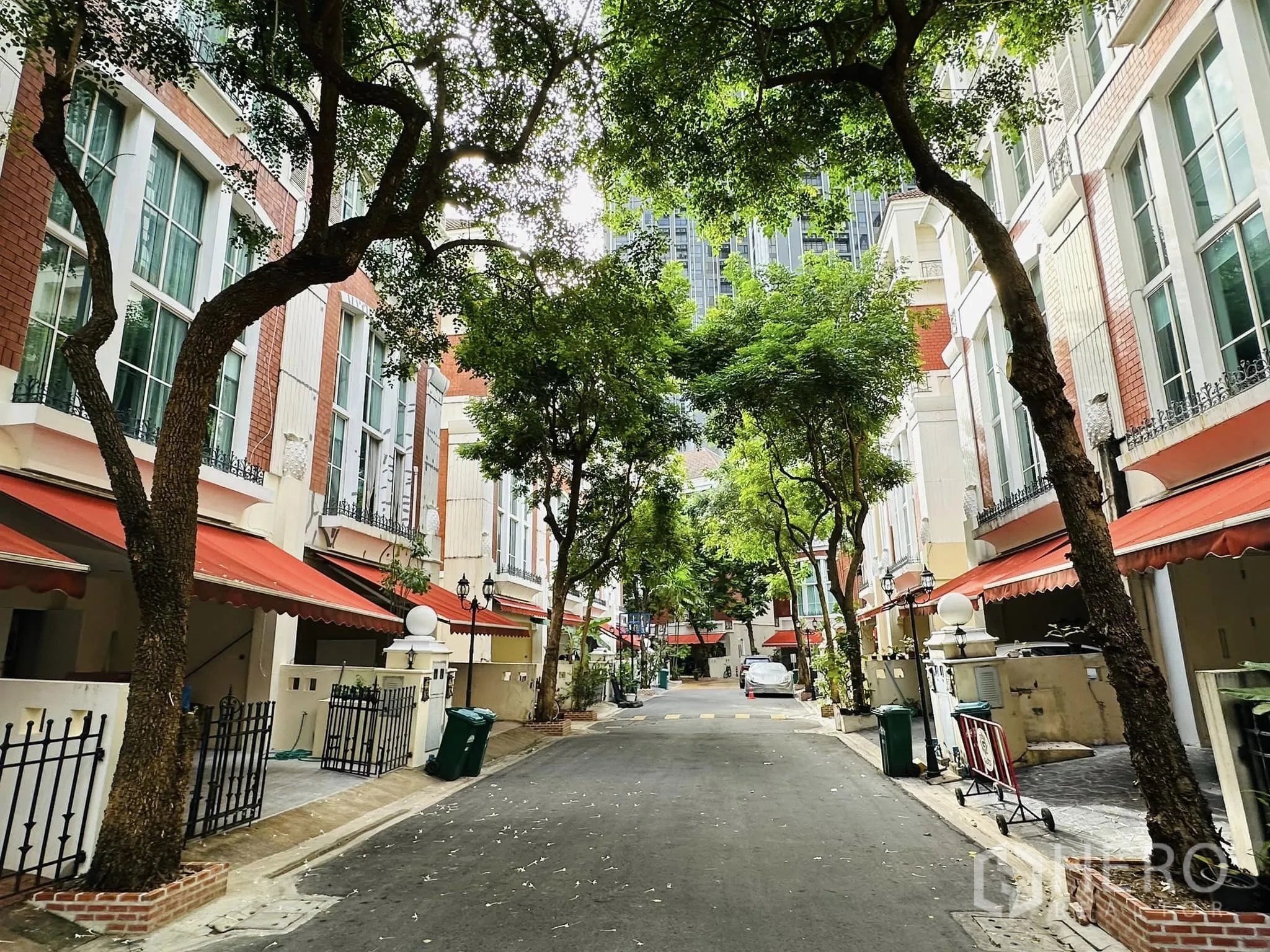 Townhouse for rent in Watthana, Bangkok - Tree-lined private lane within a townhouse compound, red awnings and gated entries on both sides.