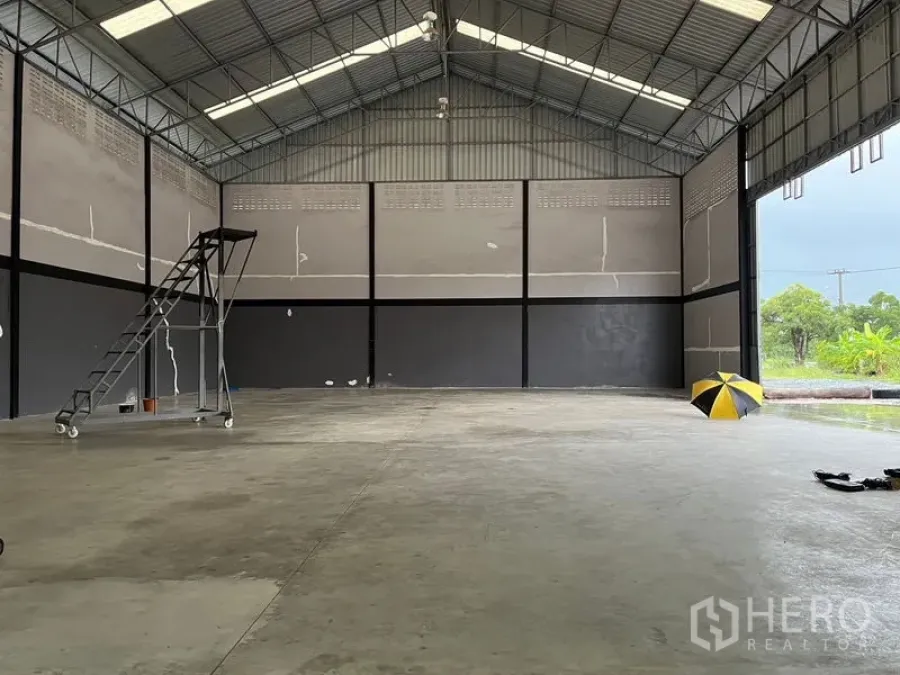 Warehouse for rent in Lat Krabang, Bangkok - Open warehouse bay with high gable roof, large roller shutter opening to greenery, and mobile ladder on the floor.