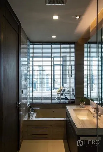 คอนโด ให้เช่า - ห้องน้ำสมัยใหม่พร้อมอ่างอาบน้ำและเคาน์เตอร์อ่างล้างหน้า มองออกไปยังหน้าต่างมีมู่ลี่