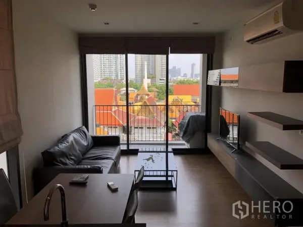 Condominium for rent in Watthana, Bangkok - Living room with sofa, TV wall, and floor-to-ceiling doors opening to a balcony with city and temple roof views.