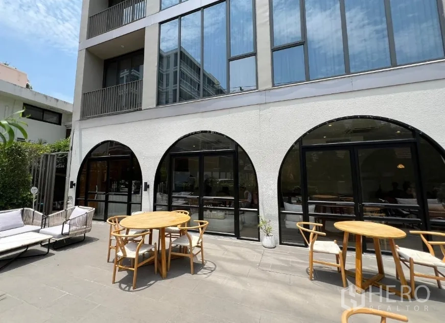 零售空间 出租在Watthana, Bangkok - 现代建筑外立面，黑色拱形玻璃门通向阳光庭院，摆放圆形木桌。