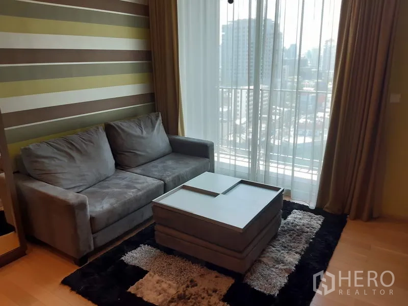 คอนโด ให้เช่าในKhlong Toei, Bangkok - ห้องนั่งเล่นสว่าง โซฟาสีเทา ผนังลาย และประตูกระจกสูงจรดเพดานชมวิวเมืองจากระเบียง