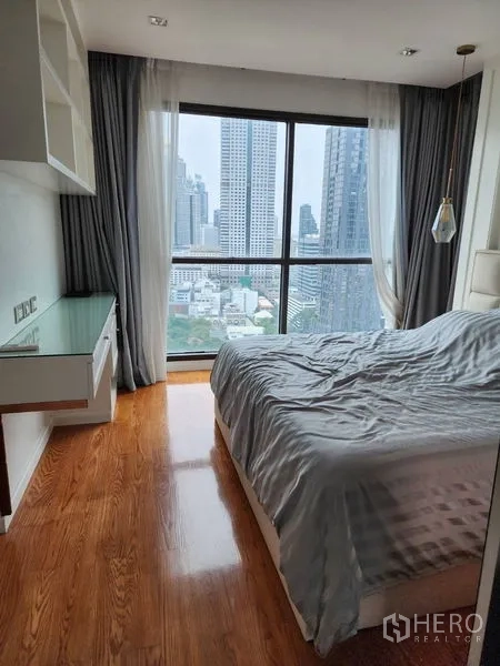 公寓 出租在Bang Rak, Bangkok - 卧室角落设落地窗，城市景观，带内置书桌与灰色窗帘。