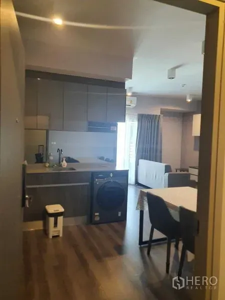 Condominium for rent - Open-plan kitchen with grey cabinets, peninsula counter and in-unit washer leading to a bright living area with balcony curtains.
