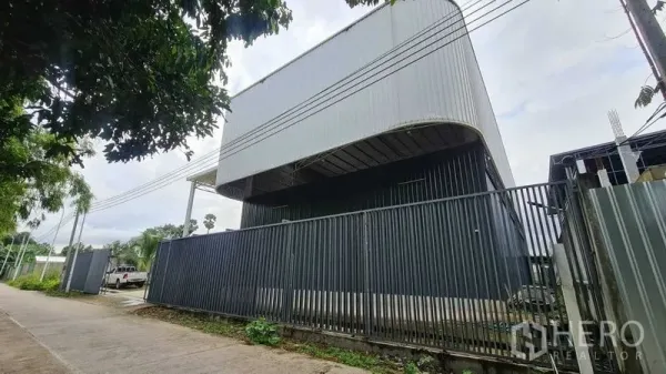Warehouse for rent in Mueang Nonthaburi, Nonthaburi - Modern warehouse exterior with a curved metal roof and secure fence seen from the street.