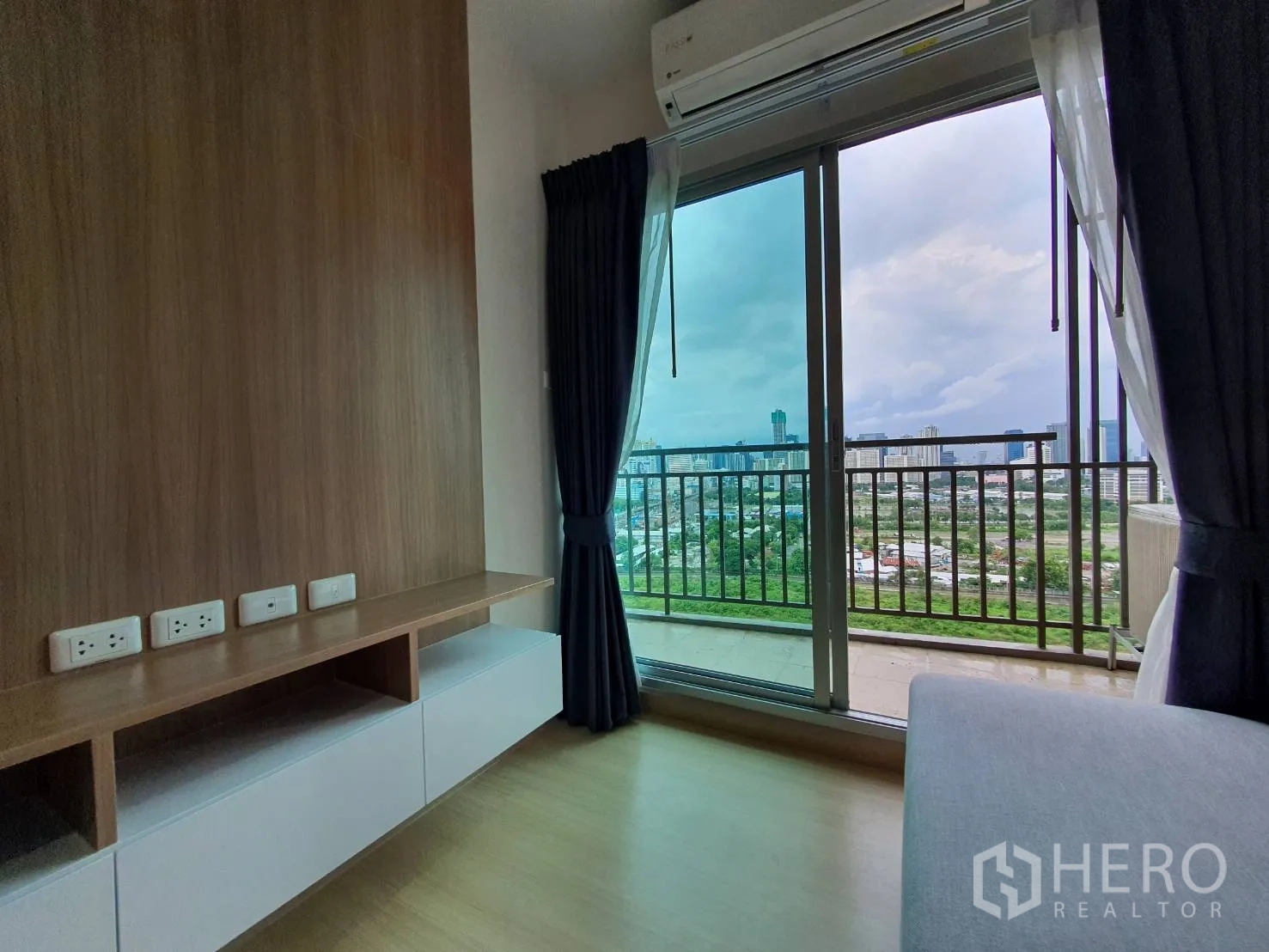 คอนโด for rent in Huai Khwang, Bangkok - ชั้นวางทีวีผนังไม้ติดประตูกระจกบานเลื่อนออกระเบียง เห็นวิวสกายไลน์และพื้นที่สีเขียว มีเครื่องปรับอากาศ