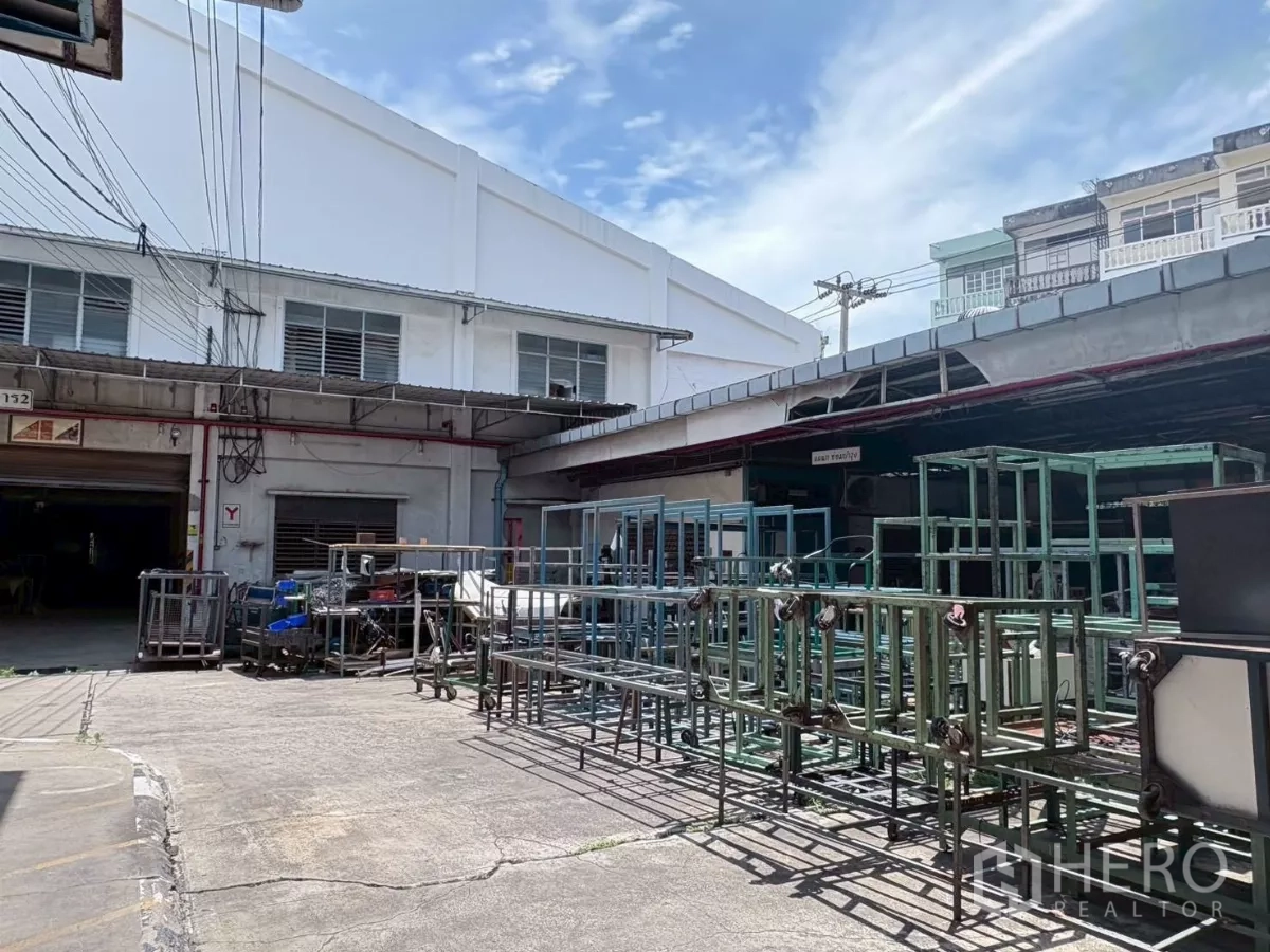 工厂 出租或出售在Bang Khun Thian, Bangkok - 混凝土院场，摆放金属货架，周围为车间建筑。