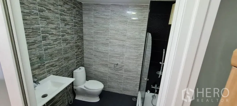 Townhouse for rent in Watthana, Bangkok - Modern bathroom with gray stone-look tiles, toilet, vanity, and glass shower.