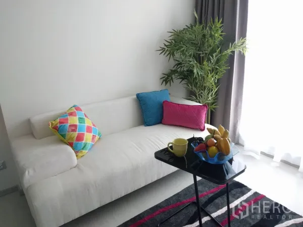 Condominium for rent in Khlong Toei, Bangkok - Bright lounge with white sofa, colorful pillows, indoor plant, and floor-to-ceiling window.