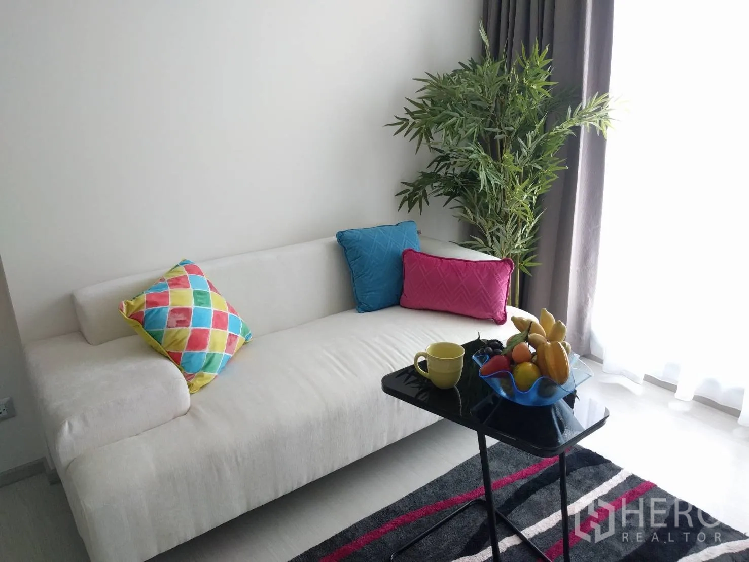 Condominium for rent in Khlong Toei, Bangkok - Bright lounge with white sofa, colorful pillows, indoor plant, and floor-to-ceiling window.