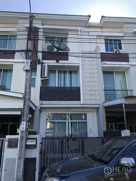 Townhouse for rent in Yan Nawa, Bangkok - Three-storey modern townhouse exterior in Yan Nawa, Bangkok, with balconies, sliding glass doors, satellite dish, air-conditioning units, gated parking and overhead cables on a sunny day.