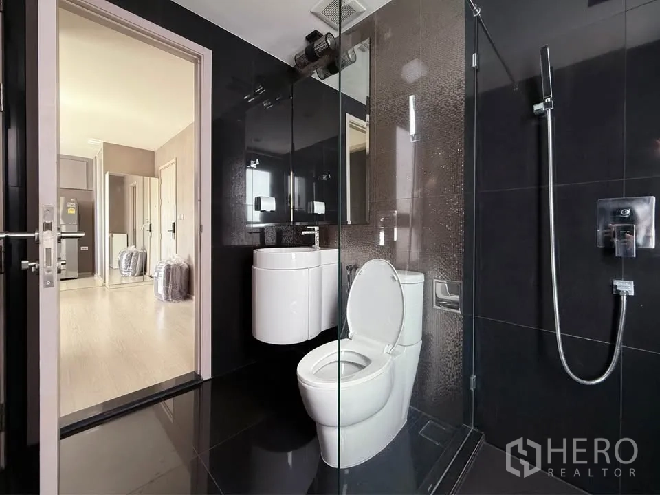 Condominium for rent in Khlong Toei, Bangkok - Sleek black-tiled bathroom with glass shower, toilet and curved vanity opening to the hallway.