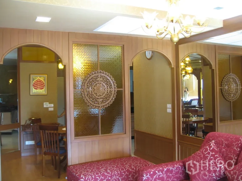 Condominium for rent in Khlong Toei, Bangkok - Living area with warm wood paneling, arched mirrors, decorative frosted glass and a chandelier beside a red chaise.