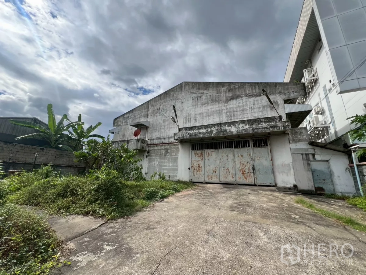 คลังสินค้า ให้เช่าในPhra Khanong, Bangkok - ภาพภายนอกโกดังคอนกรีตเก่า ประตูเหล็กบานเลื่อนเป็นสนิม พร้อมทางลาดรถและพื้นที่สีเขียว ใต้ท้องฟ้ามีเมฆในเขตพระโขนง กรุงเทพฯ
