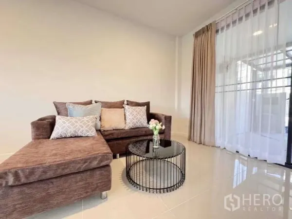 Townhouse for rent in Min Buri, Bangkok - Bright living room with brown sectional sofa, round glass coffee table, and sheer curtains by sliding door.