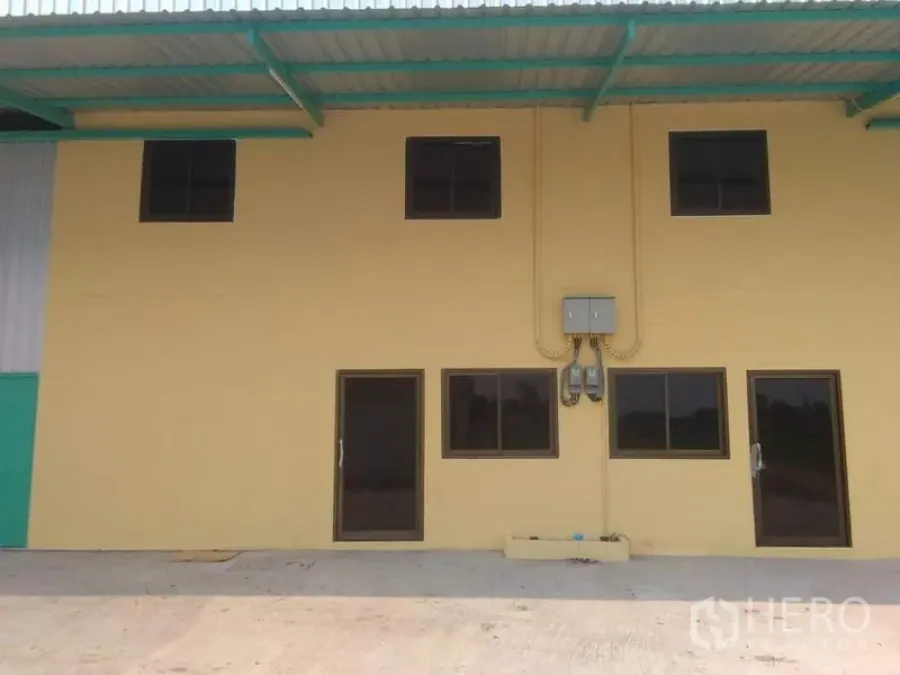 Factory for rent in Khlong Luang, Pathum Thani - Two-story factory office facade with yellow walls, dark-framed windows, electrical meters and a covered loading canopy on a concrete apron in Khlong Luang.