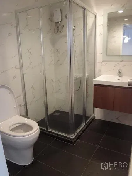 公寓 出租在Bang Rak, Bangkok - 现代卫生间，玻璃淋浴间与热水器，带镜洗手台，大理石纹墙面与深色地砖。