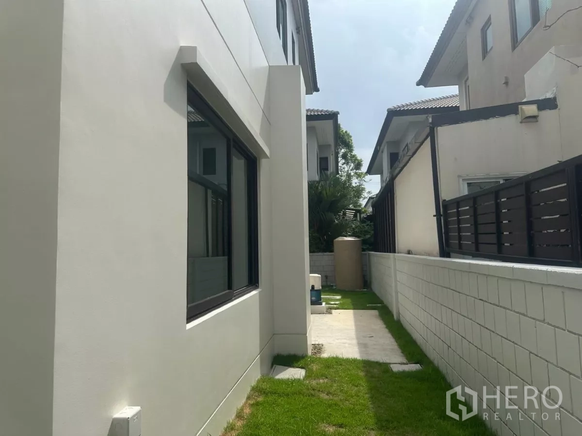 Detached House for sale in Samut Prakan - Narrow service side yard with grass, concrete path and boundary wall behind the house.