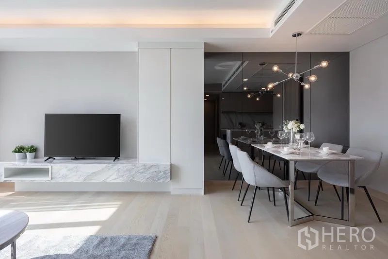 Condominium for rent in Khlong Toei, Bangkok - Modern dining space with designer light fixture beside a wall-mounted TV on a marble console and mirrored feature wall.